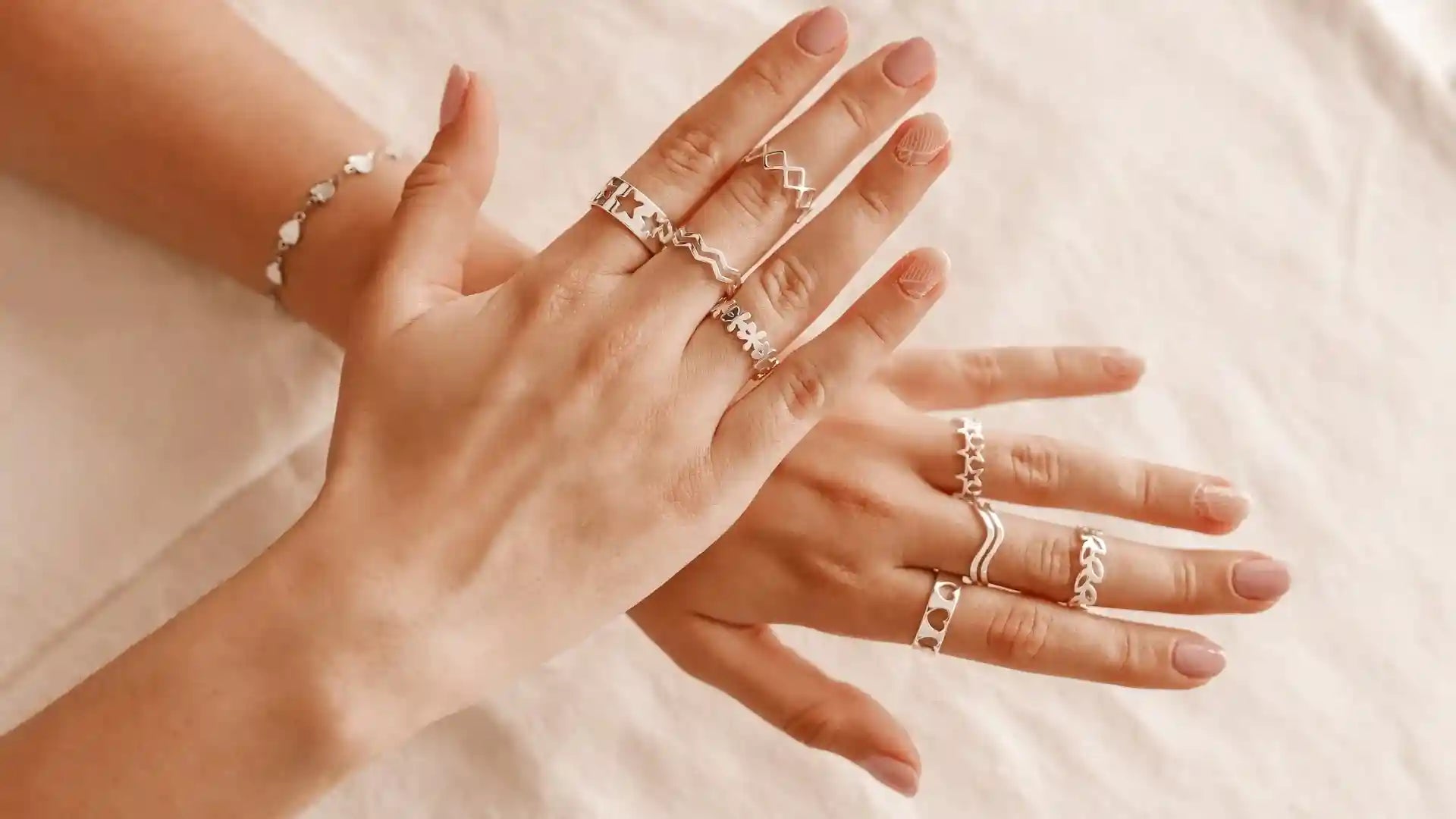 Close-up of hands wearing multiple silver rings with geometric and floral designs, styled with a delicate bracelet on a neutral fabric background.